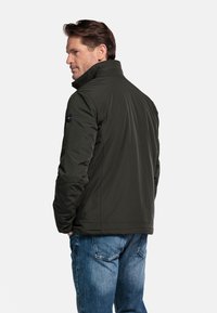 Donkergroene gewatteerde jas met een hoge kraag en een zwart cirkelvormig logo op de linkerschouder. Draagt over een spijkerbroek, met een eenvoudig ontwerp.