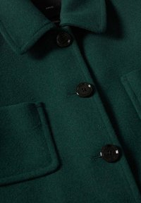 Gros plan sur un manteau en laine vert foncé montrant des boutons noirs, un col et deux poches avant sur un fond de tissu lisse.