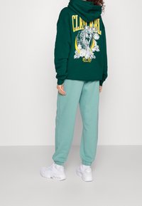 Sudadera verde de gran tamaño con gráfico floral y texto amarillo, combinada con pantalones jogger azul claro y zapatillas deportivas blancas.
