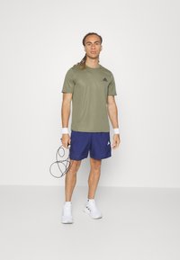 Olive green athletic t-shirt, blue shorts, white sneakers. Model holds a jump rope; features logo accents on clothing. Minimalist background.