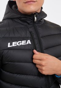 Svart pufferjacka med dragkedja, med hög krage och kviltad textur. Logotypen "LEGEA" i vitt på framsidan.