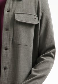 Grijze gestructureerde overhemden met een enkele borstzak en een zichtbaar logo-detail. Draagt over een bordeauxrood shirt, wat contrasterende kleuren laat zien.