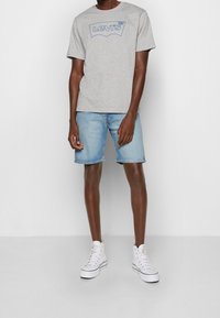 Harmaa puuvillapaita, jossa on sininen Levi's-logo, yhdistettynä vaaleansiniin farkkushortsit. Mallilla on päällä valkoiset korkeakärkiset lenkkarit ja hän seisoo neutraalilla taustalla.