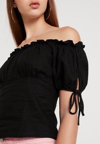 Top nero off-the-shoulder con maniche a sbuffo, bordo arricciato elasticizzato e dettagli con lacci alle aperture delle maniche. Tessuto di superficie liscia.