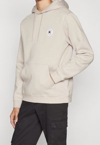 Ljusbeige hoodie med framficka, dragsko-kappa och cirkulär logotyp med en blå stjärna. Mjuk tyg med ribbade ärmar och nederkant.
