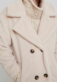 Dubbelknäppt, mjukt, ljusrosa kappa med en plyschstruktur, stora slag och svarta knappar, bärs över en strukturerad beige tröja.