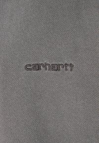 Tela gris con una textura suave que presenta un logotipo bordado de "carhartt" en un tono más oscuro. La vista en primer plano resalta el detalle del logotipo.
