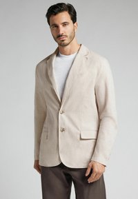 Blazer de lino beige claro con solapa de muesca, dos botones y bolsillos frontales. Llevado sobre una camisa gris claro, con una textura suave.