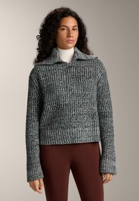 Šedý pletený pulovr se zipem u krku, s vroubkovanou strukturou a širokými rukávy, doplněný hnědými legínami.