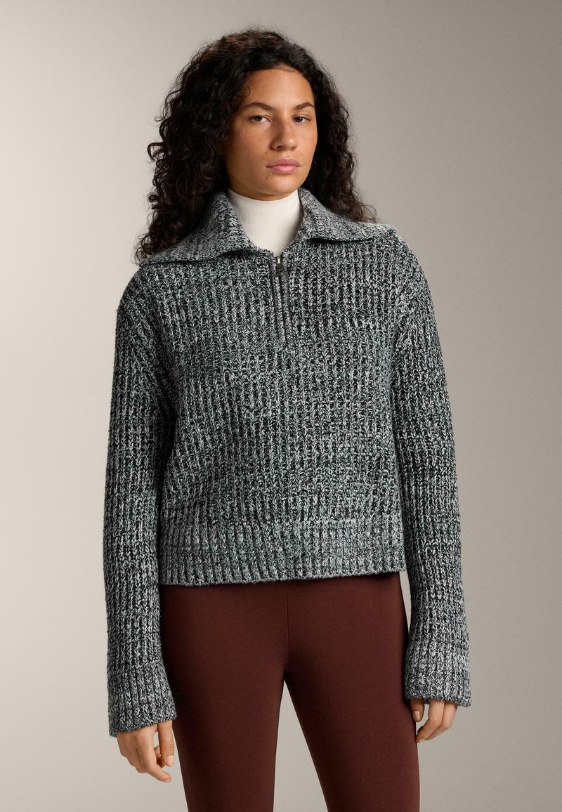 Šedý pletený pulovr se zipem u krku, s vroubkovanou strukturou a širokými rukávy, doplněný hnědými legínami.