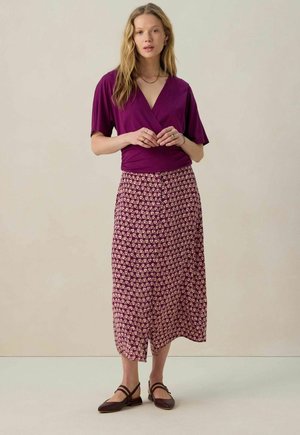 Haut en violet à manches courtes associé à une jupe longue à motifs dans des teintes de violet et de crème. Des ballerines marron complètent la tenue.