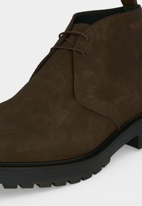 Bottine en daim marron avec semelle noire épaisse, lacets à l’avant et détails de couture visibles sur un fond uni clair.