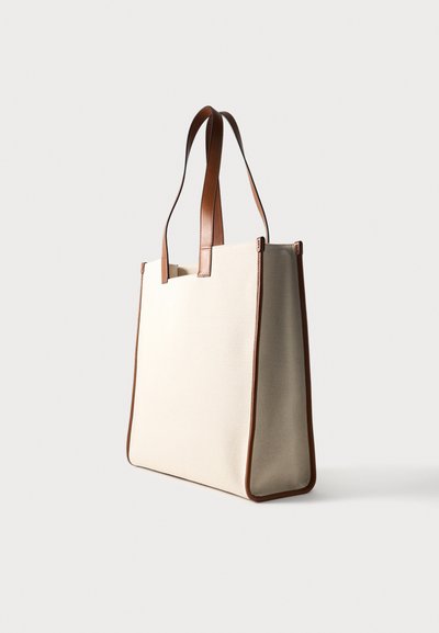 Μπεζ ορθογώνια τσάντα tote με καφέ δερμάτινα χερούλια και διακοσμητικά, όρθια σε απλό ανοιχτό φόντο.