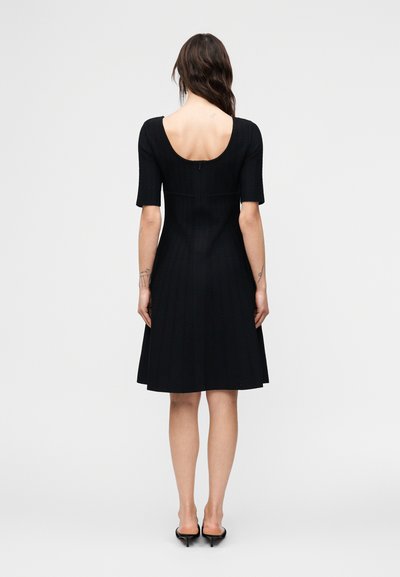 Γυναίκα με κυματιστά καστανά μαλλιά που φοράει μαύρο φόρεμα μήκους μέχρι τα γόνατα με μανίκια ως τον αγκώνα και ανοιχτή πλάτη, στέκεται γυρισμένη με την πλάτη, φορώντας μαύρες γόβες.