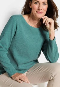 Frau sitzt, trägt einen strukturierten, türkisfarbenen Langarmoberteil und beigen Hosen, ruht ihr Kinn auf der rechten Hand und die linke Hand auf dem Knie, lächelt leicht.