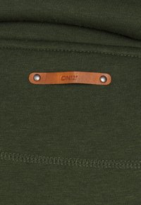 Kakigrön tyg med en slät textur; har en brun läderetikett med "ONLY" präglat och silvernitar. Synliga sömmar.