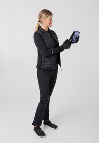 Schwarze isolierte Weste über einem Langarmoberteil, kombiniert mit schwarzen Hosen und Handschuhen, während sie ein Smartphone mit einer Berganzeige hält.