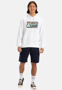 Sudadera blanca con un diseño gráfico que presenta un patrón y logo en verde y negro, combinada con pantalones cortos azul marino y zapatillas negras.