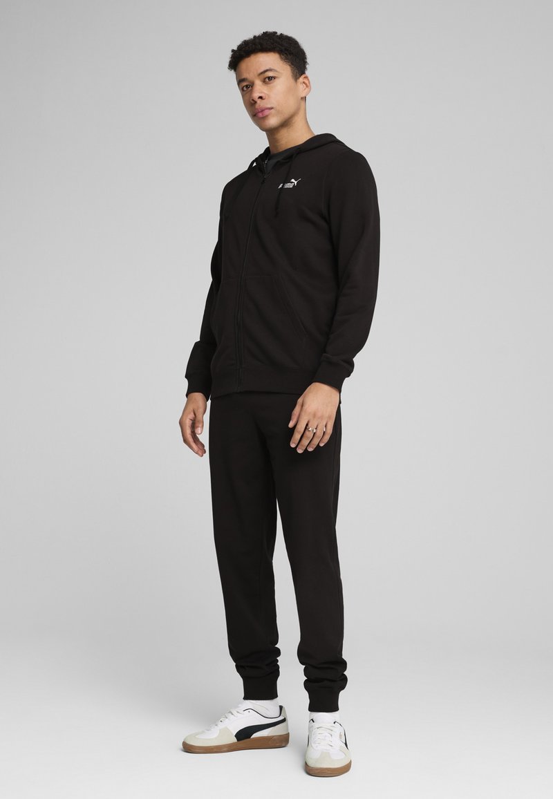 Sort zip-up hoodie i blødt stof med frontlommer, parret med matchende sorte joggers og hvide sneakers med sorte accenter.