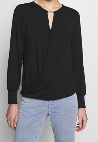 Haut noir avec un décolleté rond, détail en goutte d'eau et devant drapé. Manches longues avec des poignets ajustés, fabriqué dans un tissu doux et lisse.