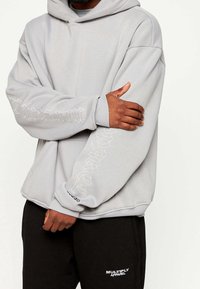 Světle šedá mikina s velkou kapucí a vyšitým textem na rukávech. V kombinaci s černými tepláky, které mají bílé logo na stehnu.