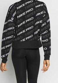 Felpa nera Nike Pro con stampa del logo bianco ben in evidenza, design cropped, polsini a coste e una texture liscia. Abbinata a leggings neri.