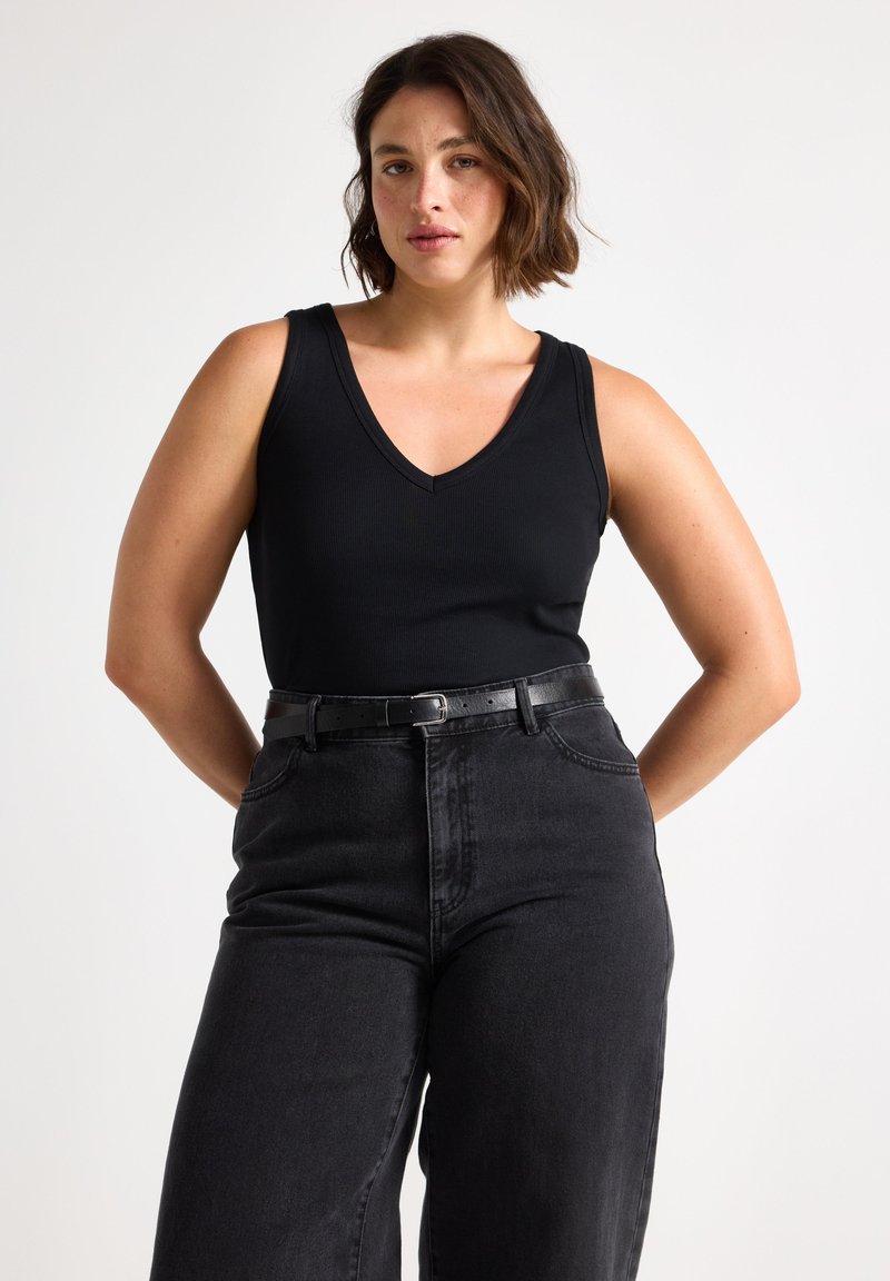 Femme aux cheveux courts et bruns portant un débardeur noir moulant à col en V, rentré dans un jean noir taille haute avec une ceinture noire.
