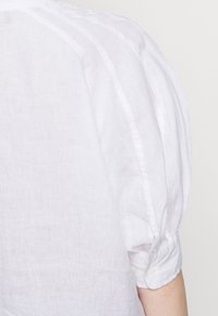 Chemise blanche en lin à manches longues avec manches légèrement bouffantes et poignets boutonnés, portée sur le bras et le dos d'une personne.