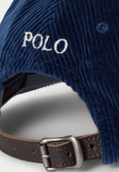 Σκούρο μπλε καπέλο από κοτλέ με κεντητό λογότυπο "POLO". Διαθέτει καφέ δερμάτινο λουρί με ατσάλινη αγκράφα. Υφασμάτινη υφή και καμπυλωτή γείωση.