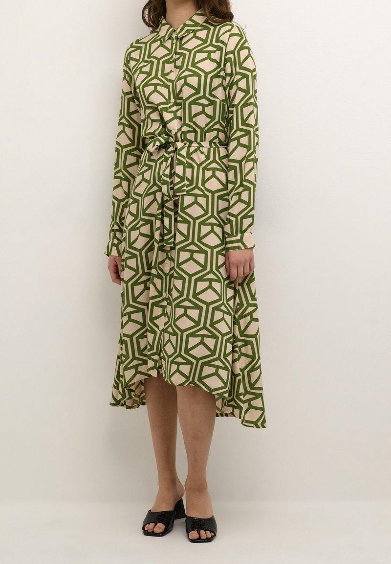 Femme portant une robe beige à manches longues avec des motifs géométriques verts, une ceinture nouée à la taille et des sandales à talons ouvertes noires.