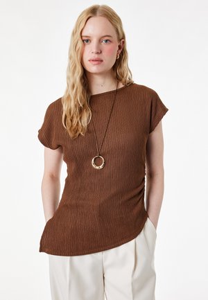 Jonge vrouw met lang blond haar, gekleed in een bruin geribd kort mouwtopje, crèmekleurige broek en een lange ketting met een cirkelvormige hanger.
