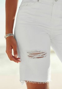 Un bras bronzé orné de bracelets en perles à côté de shorts en denim blanc présentant une déchirure usée et un ourlet effiloché sur la cuisse.