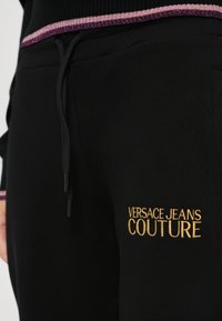 Sorte joggebukser med gull "VERSACE JEANS COUTURE" broderi, med snor og en teksturert midje i en kontrastfarge.