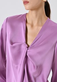 Primo piano di una donna che indossa una blusa lucida color lilla con un dettaglio a nodo attorcigliato sul davanti e orecchini quadrati in oro, capelli raccolti.