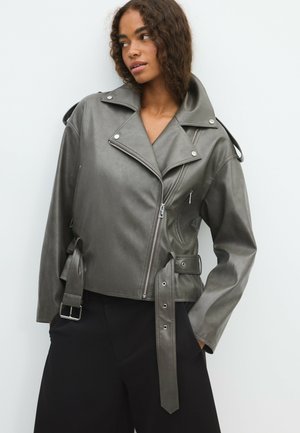 Jeune femme portant une veste de motard en cuir gris avec une fermeture éclair asymétrique et une ceinture à la taille, debout avec les mains dans les poches d’un pantalon noir à jambes larges.
