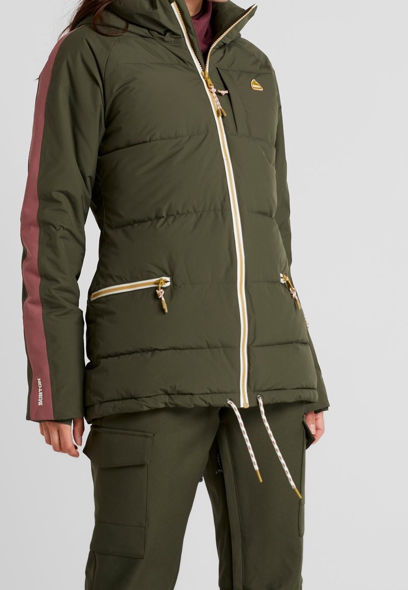 En person som bär en grön puffjacka med rosa accenter, tillsammans med matchande gröna byxor, står mot en enkel bakgrund.