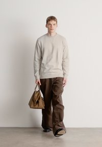 Μπεζ πουλόβερ και χαλαρό καφέ παντελόνι, κρατώντας μια μικρή καφέ τσάντα. Ομαλές υφές, άνετη εφαρμογή. Το μοντέλο περπατά προς τα εμπρός μπροστά σε απλό φόντο.