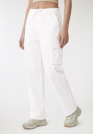 Kobieta ubrana w białe spodnie typu cargo z bocznymi kieszeniami, beżowy krótki top i jasnoszare trampki, stojąca na jednolitym białym tle.