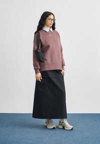 Γυναίκα με μακριά μαλλιά και γυαλιά, που φοράει πουλόβερ σε μοβ απόχρωση, μαύρη μακριά φούστα, μπεζ αθλητικά παπούτσια και μια μικρή μαύρη τσάντα ώμου.