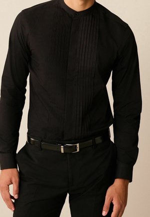 Homme portant une chemise noire ajustée à col mandarin avec un détail plissé à l'avant et un pantalon noir maintenu par une ceinture en cuir noire avec une boucle argentée.