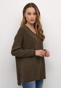 Brun ribbknittröja med V-ringning, långa ärmar och sidoflikar. Texturerad tyg och avslappnad passform. Buren över blå jeans.