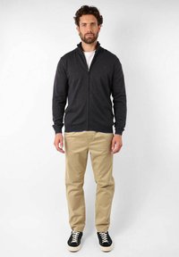 Veste zippée gris foncé avec un col haut, associée à un pantalon chino beige et des baskets noires avec des lacets blancs. Texture lisse et design ajusté.