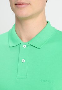 Grön pikétröja i texturerat tyg. Har en klassisk krage, tre knappar och en diskret "Esprit"-logotyp broderad vid fållen.