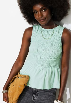 Femme aux cheveux bouclés portant un top sans manches vert clair, un pantalon gris, des bijoux en argent, et tenant un sac en daim beige.