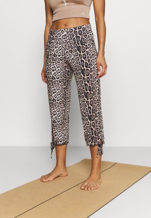 Vrouw die blootsvoets staat op een yogamat, gekleed in cropped legging met luipaardprint met trekkoorden en een beige sportbeha.