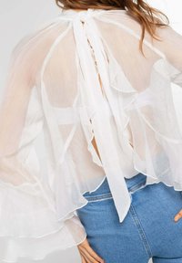 Blusa blanca transparente con mangas con volantes y cierre de lazo en la parte posterior. Tejido suave y ligero con una textura translúcida.