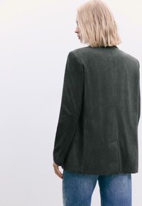 Mørkegrøn ruskindsblazer med en glat overflade, enkelt ventil bagpå og en skræddersyet pasform, båret over lyseblå denimjeans.