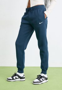 Tummansiniset Nike-housut, joissa on elastinen vyötärö ja nyöri. Vasemmalla puolella on valkoinen logo. Yhdistetty mustavalkoisiin tennareihin.