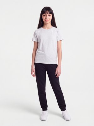 Young woman with long dark hair wearing a light grey short-sleeve t-shirt, black pants, and white sneakers standing against a white background.