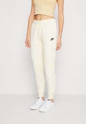 Woman in cream Nike sweatpants and white Nike sneakers standing against a plain light background.
